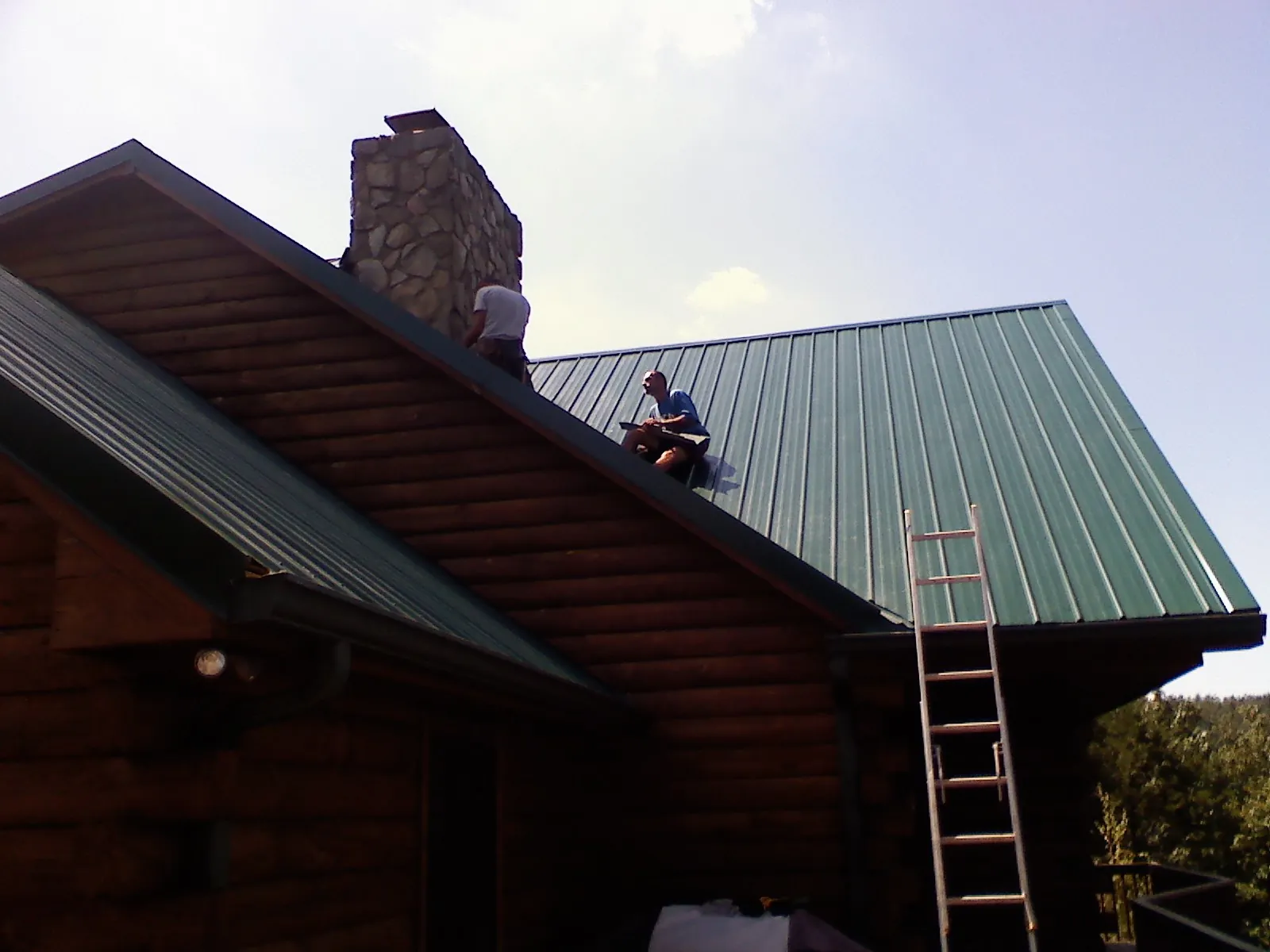 Roofers installing green metal panels beside a stone chimney on a log home in Juliustown
